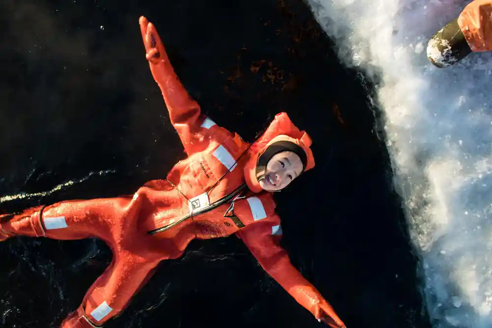 A passenger floating on her back in a survival suit, arms spread, dark water around