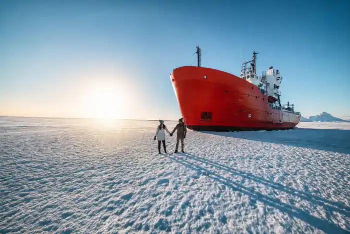 The red hull of the icebreaker standing in flat sea ice with two people on the ice below it