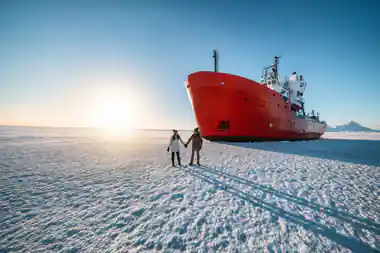 The red hull of the icebreaker standing in flat sea ice with two people on the ice below it